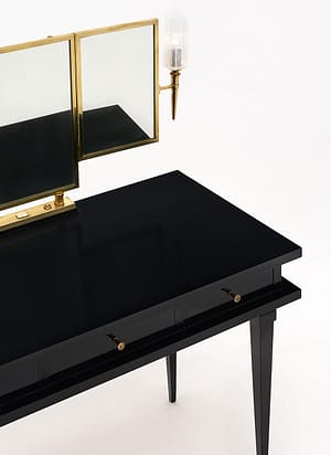 Vanity, French, made of ebonized sycamore. The vanity is in the manner of Jacques Adnet and features three dovetailed drawers and tapered legs. This elegant piece is finished in a lustrous French polish and enhanced with a triptych brass framed mirror. Each side of the mirror boasts Murano glass “torchieres” sconces that have been newly wired to fit US standards. Height to the top of the desk is 29.25”. The listed height includes the mirror.