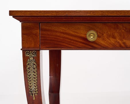 Desk, French, in the French Empire style and made of blonde figured Mahogany. This piece features four console legs that are enhanced with finely cast bronzes (stylized acanthus leaves and lion paws). Two dovetailed drawers with bronze pulls and one with original lock and working key adorn the front. The spectacular writing table sits on four ebonized bun feet and is topped with a gilt embossed tan Moroccan leather writing surface. Delve in the timeless elegance of an antique empire style desk.